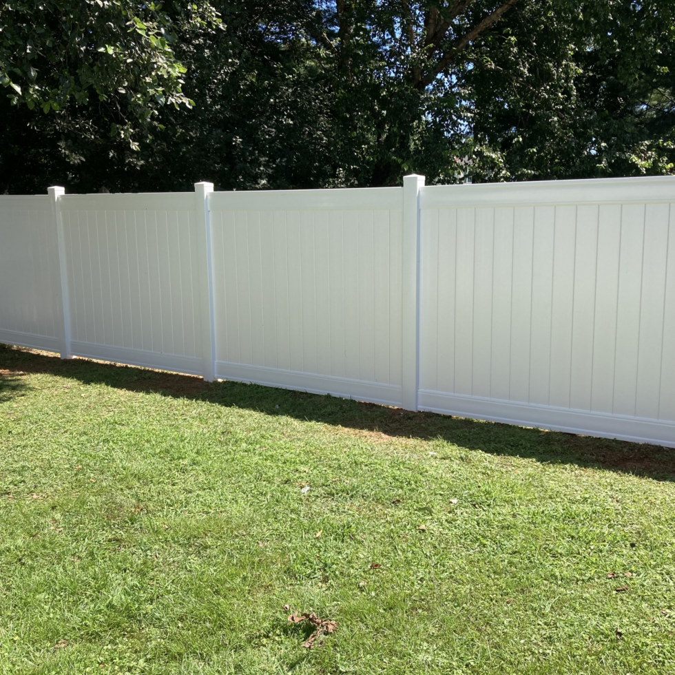 A tall, white privacy fence made of vinyl panels with vertical slats, standing along a grassy lawn. The fence has evenly spaced posts and a smooth top edge. Behind the fence, there are large, leafy trees providing shade, and the sky above is clear. The scene appears to be in a backyard or outdoor area during daytime.