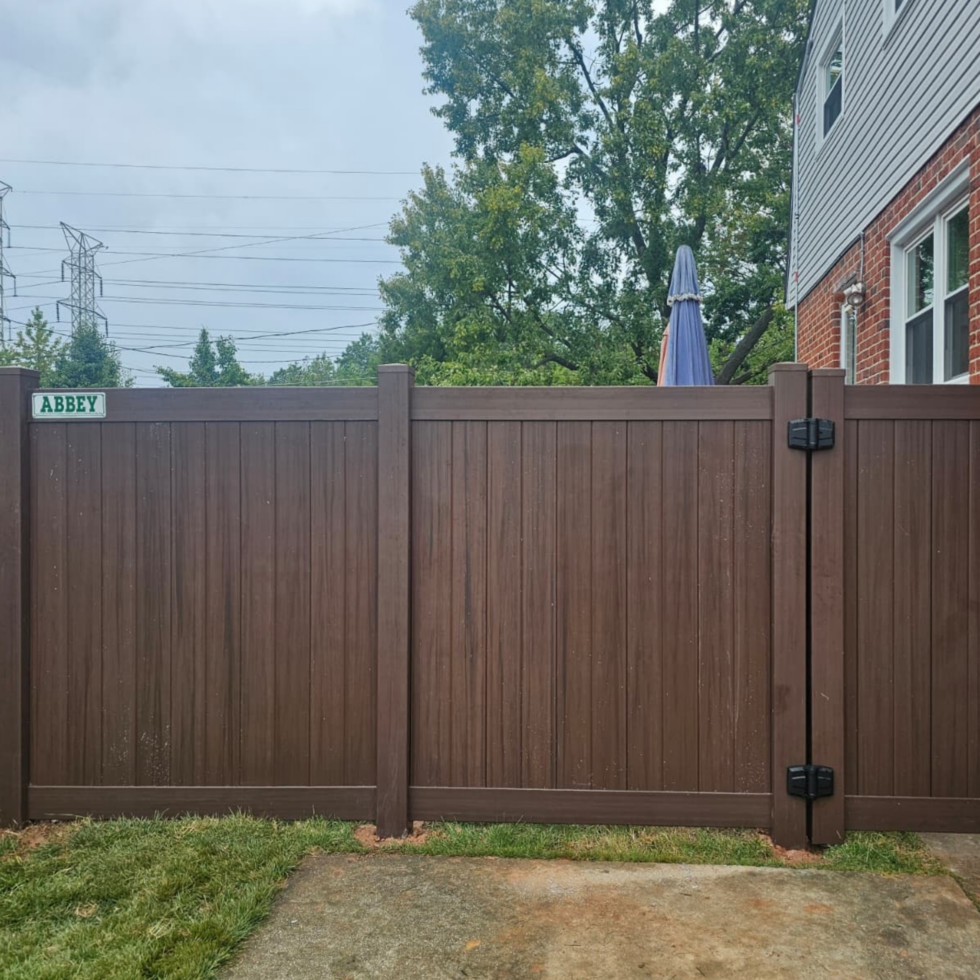 A tall brown wooden privacy fence with vertical panels and sturdy posts, situated in a backyard. There is a small green and white sign labeled "ABBEY" on the left side of the fence. Behind the fence, tall green trees and a partially visible house with gray siding and brick accents can be seen. An outdoor umbrella is also visible above the fence, and power lines are seen in the background under a cloudy sky.