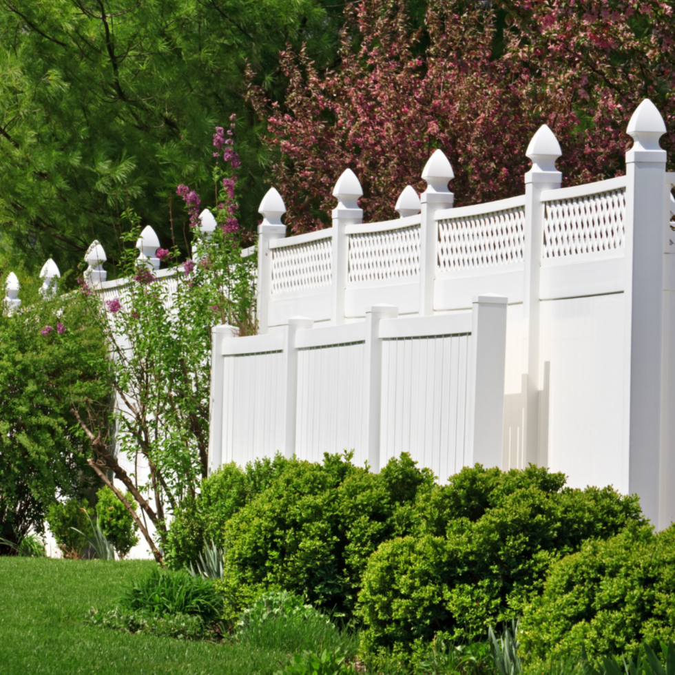 A white picket fence with decorative finials atop each post runs through a lush, green garden. Behind the fence, there are flowering bushes with pink blossoms and tall trees with dense green foliage, creating a vibrant and tranquil outdoor scene.