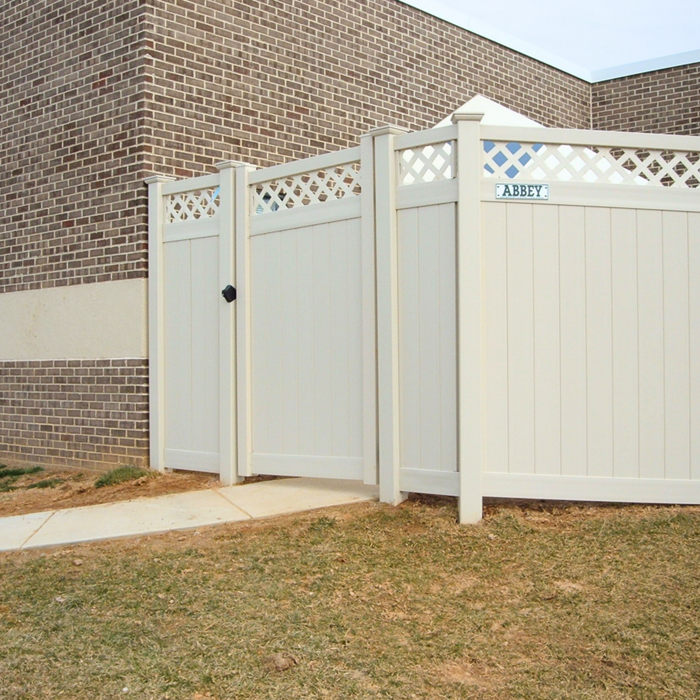 A white privacy fence with decorative lattice top panels surrounds a yard next to a brick building. There is a small gate with a black latch, and a sign on the fence that reads "ABBEY." The ground in front of the fence is a mix of grass and dirt, with a concrete sidewalk leading up to the gate. The sky above is partly cloudy.