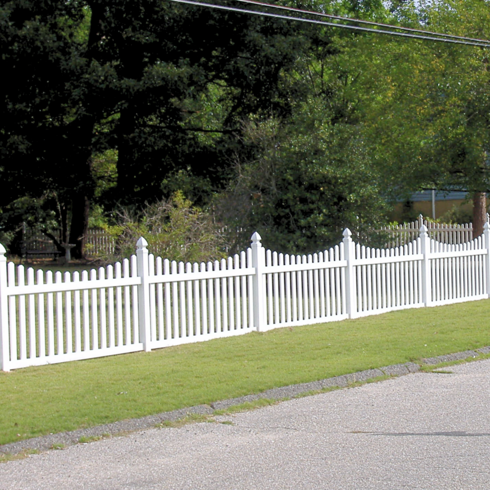 A white picket fence with decorative finials runs along a well-maintained grassy area adjacent to a paved road. Behind the fence, there are tall trees with dense green foliage, creating a lush backdrop. The scene appears to be in a suburban or rural setting during daytime.