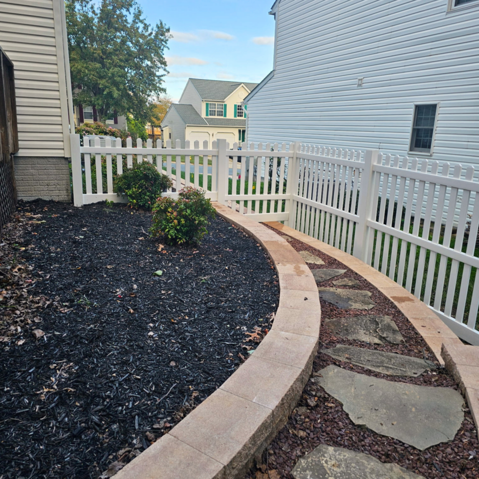 A curved stone pathway bordered by a low brick wall, running alongside a white picket fence. The pathway is set in a backyard with a garden bed covered in black mulch, containing small bushes. In the background, there are neighboring houses with light-colored siding and a partly cloudy sky above.