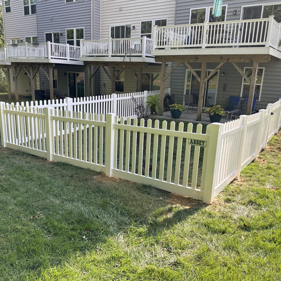 A white picket fence surrounds a small backyard area in front of a multi-story house with balconies. The fence has a gate with a small sign that reads "ABBEY." Inside the fenced area, there are potted plants, outdoor chairs, and a small garden bed, creating a cozy outdoor space. The house has gray siding, and the balconies above have white railings. The grass outside the fence is green and well-maintained.
