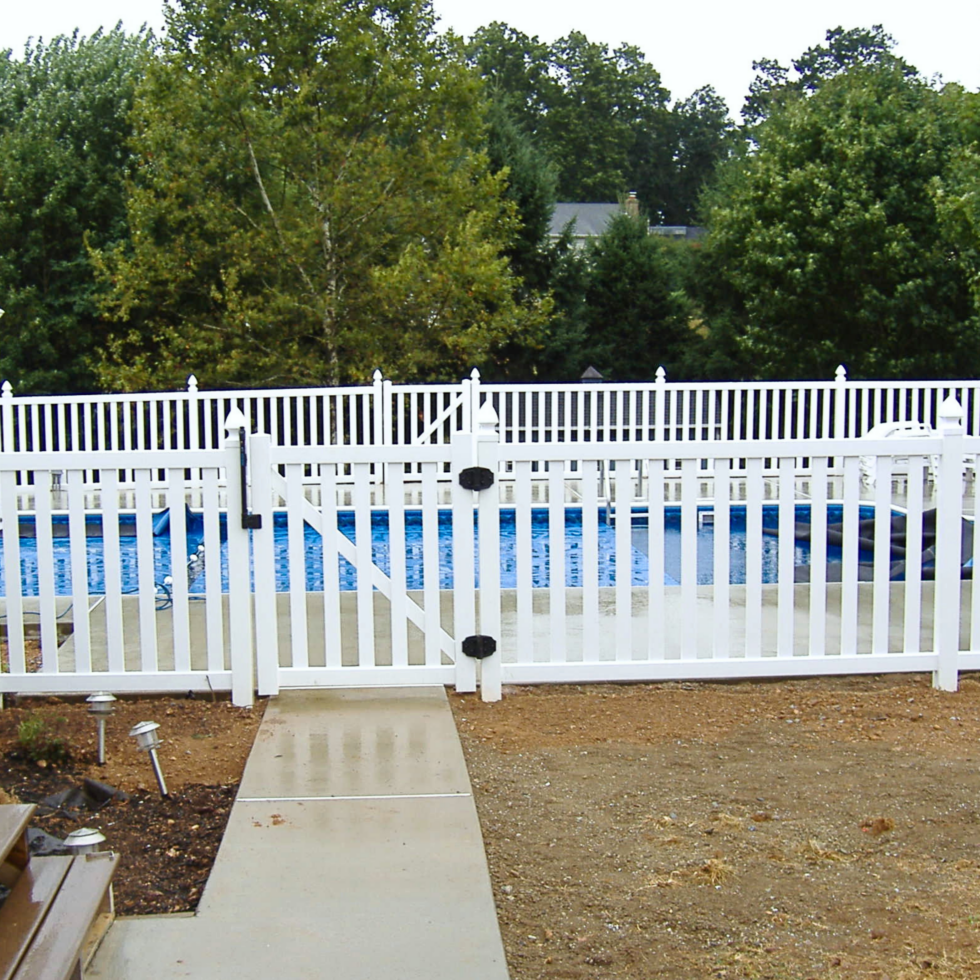A white picket fence surrounds an above-ground swimming pool, with a gate in the center. The pool is filled with blue water, and there is a concrete walkway leading to the gate. Behind the fence, there are lush green trees and some houses partially visible in the background. The ground in front of the fence is a mix of dirt and concrete.