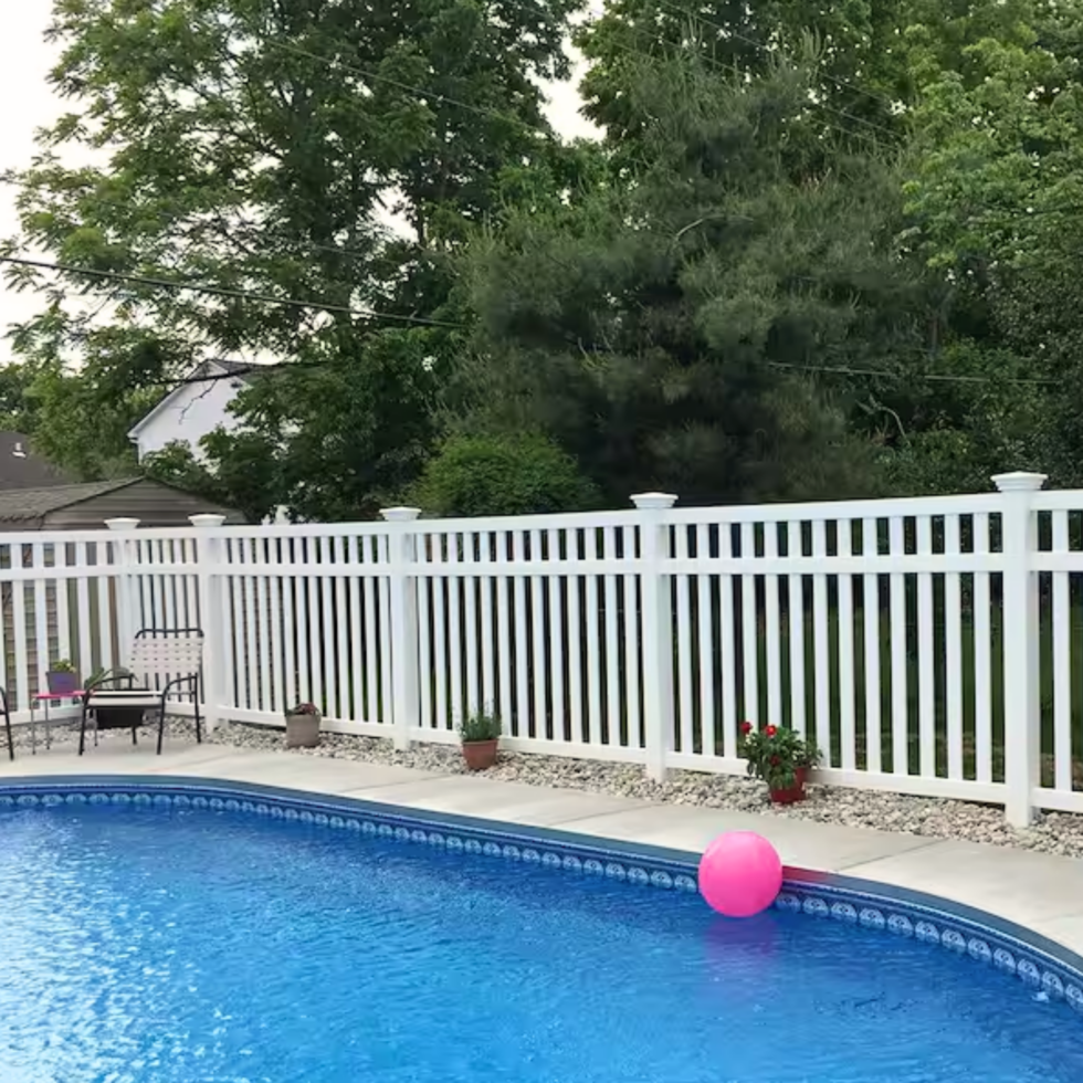 A backyard scene featuring a blue in-ground swimming pool with a pink pool float. The pool is bordered by a concrete deck with potted plants along the edge. A white picket fence surrounds the yard, and there is outdoor seating with a bench and a chair on the left side. Behind the fence, lush green trees and bushes provide a natural backdrop. The overall setting appears peaceful and inviting.