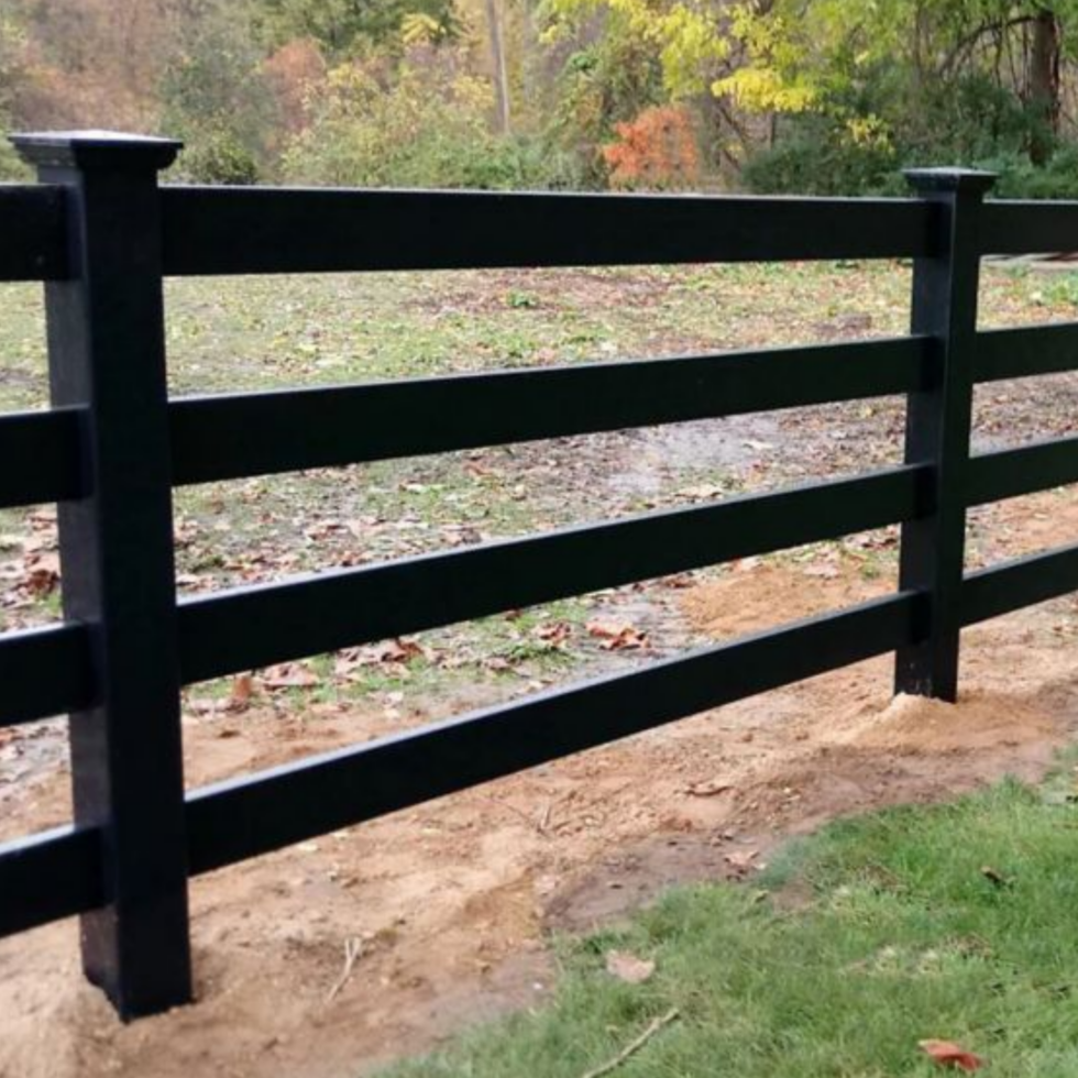 A black PVC paddock fence with vertical panels surrounds a grassy area. The fence is clean and well-maintained, providing a secure enclosure. Behind the fence, there are trees and open land, indicating a rural setting suitable for horses or livestock. The sky above is clear with a few clouds.