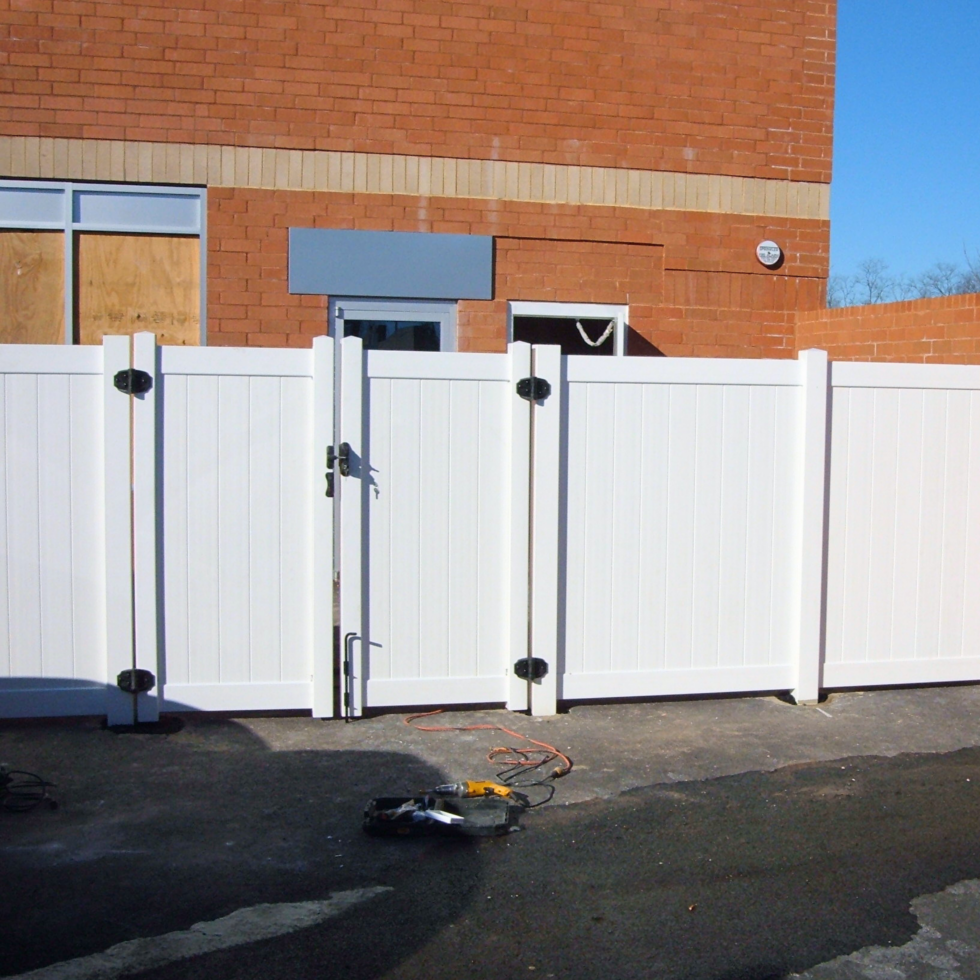 A white PVC fence with a gate is installed in front of a brick building. The gate is secured with a latch, and there are tools lying on the ground nearby, indicating ongoing installation or maintenance work. The background shows a clear blue sky.