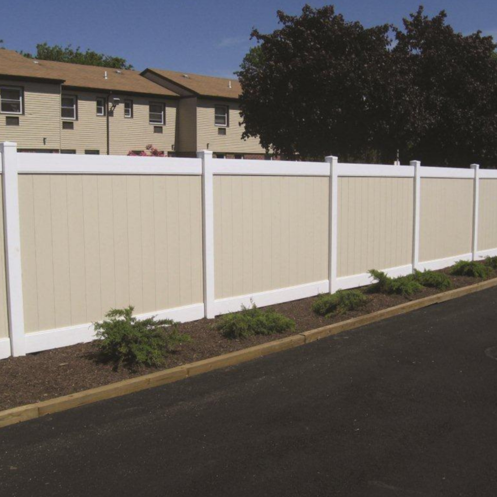 A beige vinyl privacy fence with white posts and trim, running alongside a paved surface with small landscaped plants and mulch in front of it. In the background, there are trees and multi-story residential buildings under a partly cloudy sky.