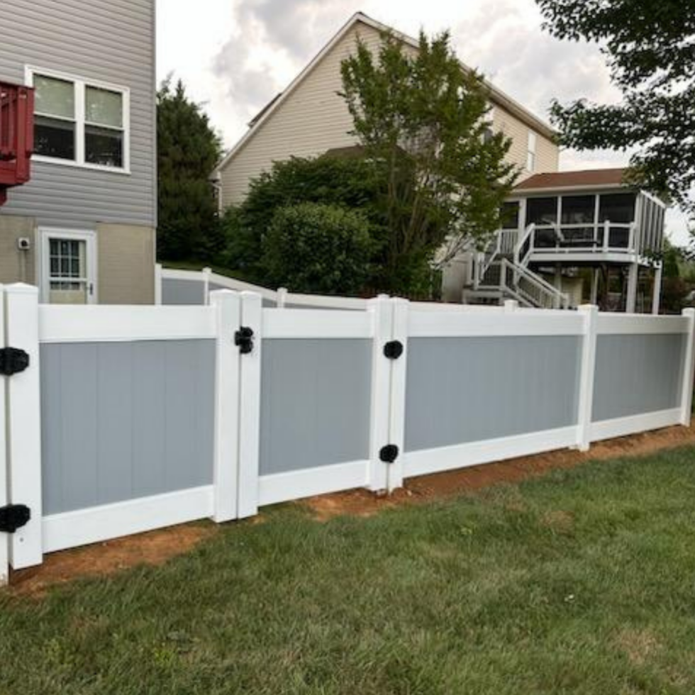 A newly installed white and gray vinyl privacy fence with black hardware, enclosing a backyard with a grassy lawn. In the background, there are two houses, trees, and a covered porch or deck. The sky is partly cloudy.