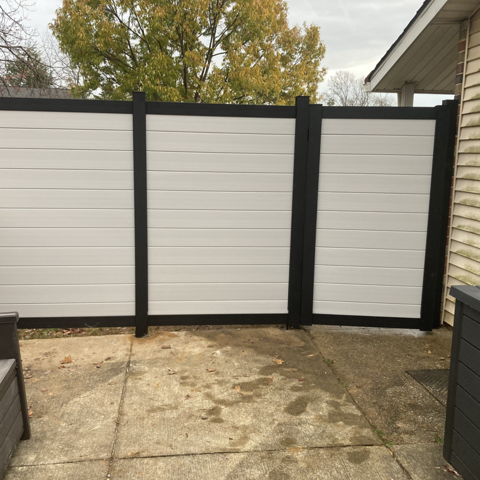 A newly installed white vinyl privacy fence with black posts and trim, situated on a concrete patio. Part of a house with beige siding is visible on the right, and a tree with autumn-colored leaves is in the background. The sky is overcast.